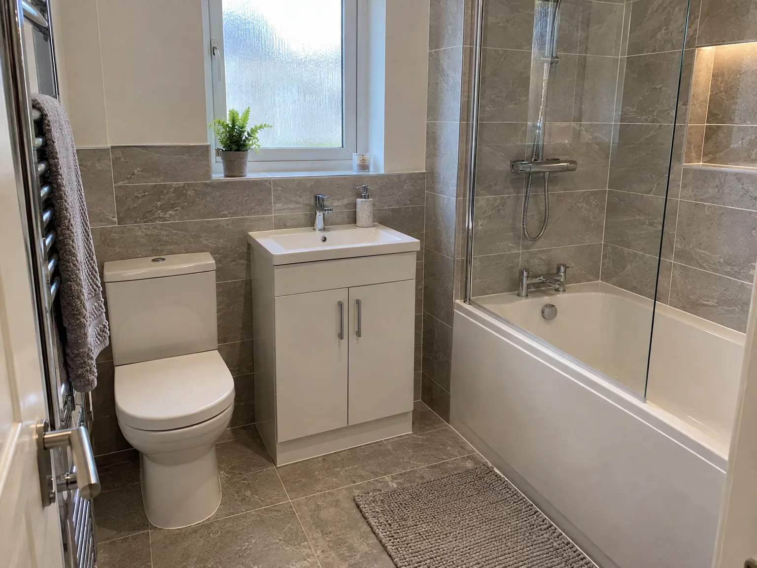 Modern bathroom with grey tile walls, beige vanity unit, toilet, bath and shower screen