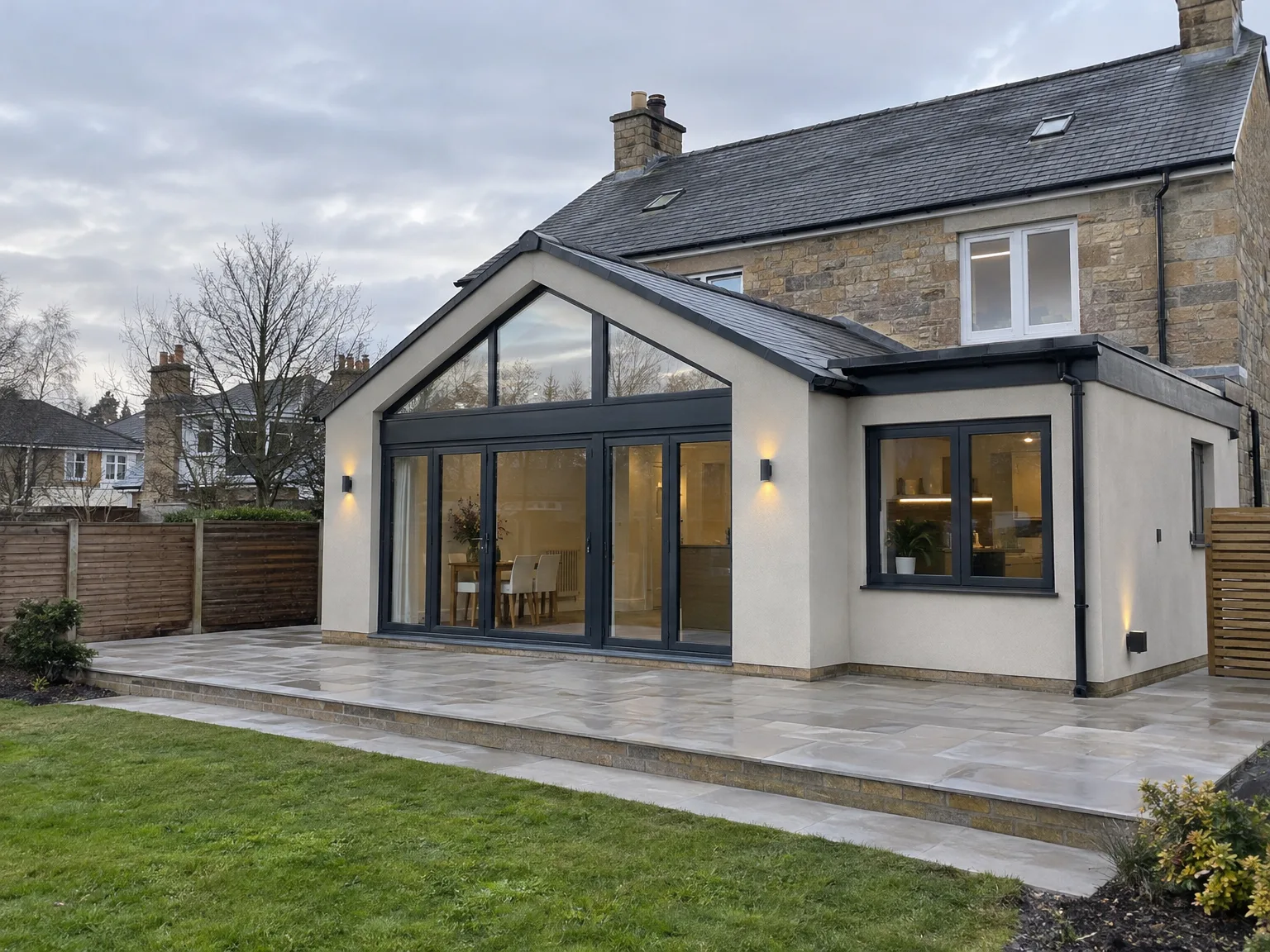 Modern house extension with white walls, large black-framed windows and stone patio adjacent to stone cottage