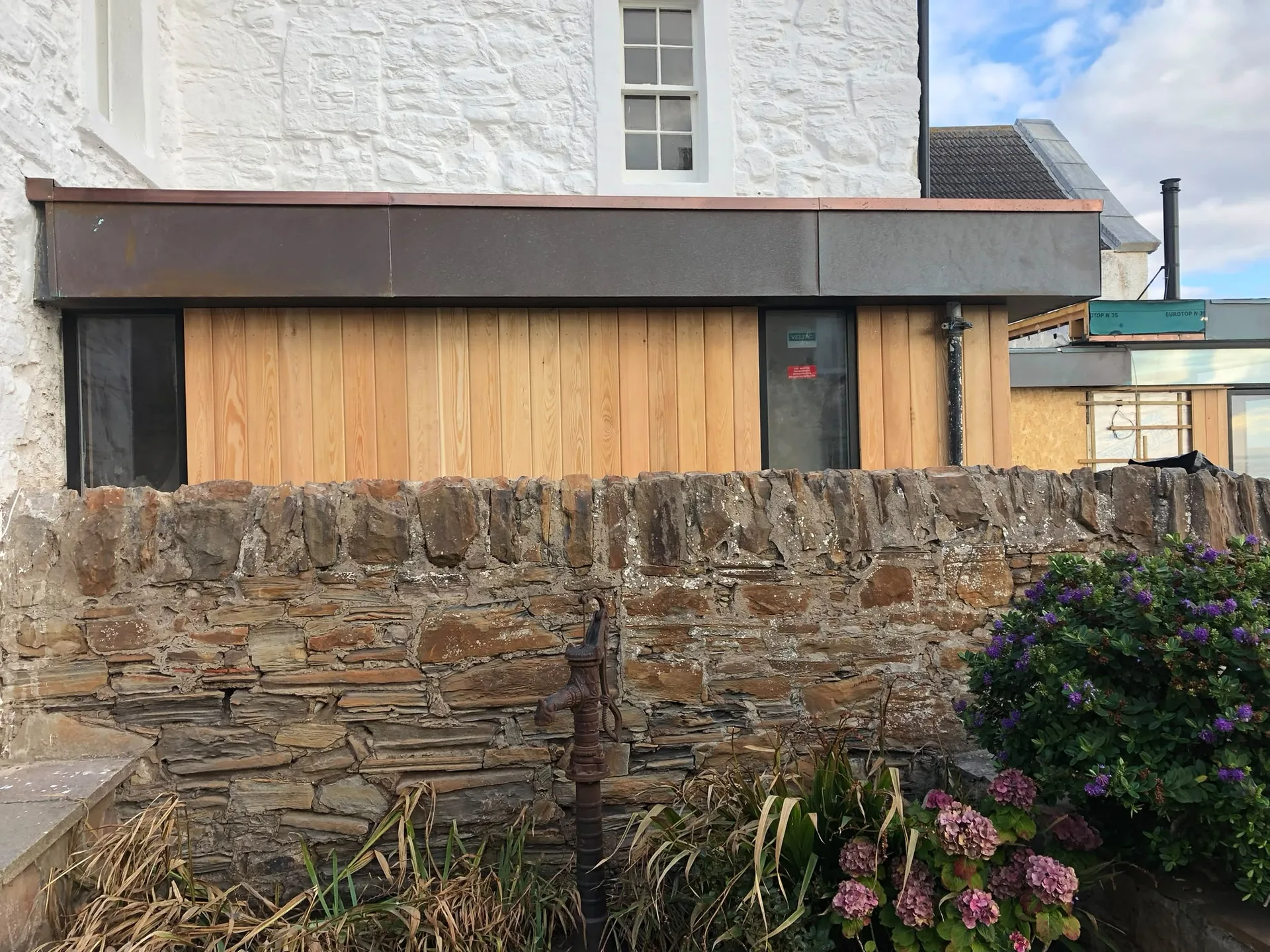 Modern timber-clad house extension with wooden facade and dark entrance doors, attached to whitewashed stone cottage with traditional pitched roof