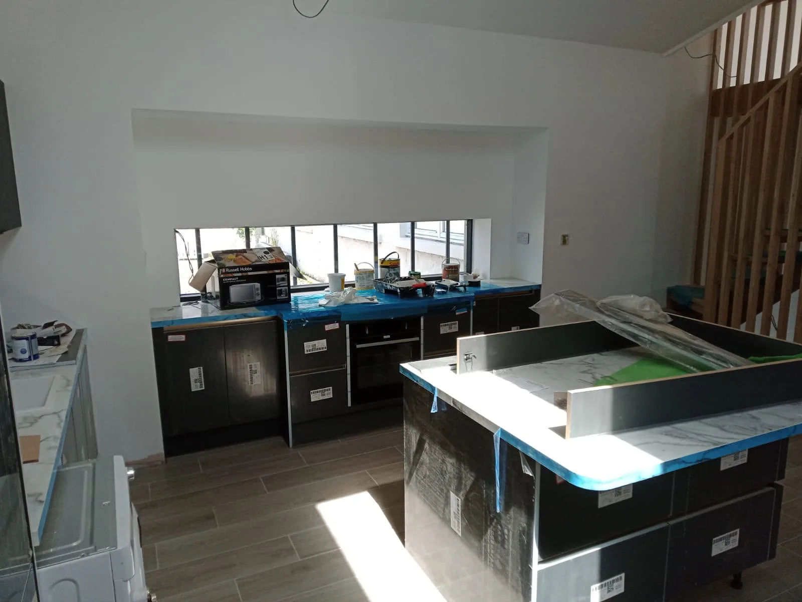 Modern black kitchen units with blue-edged counters in home renovation space with white walls and large windows providing natural light