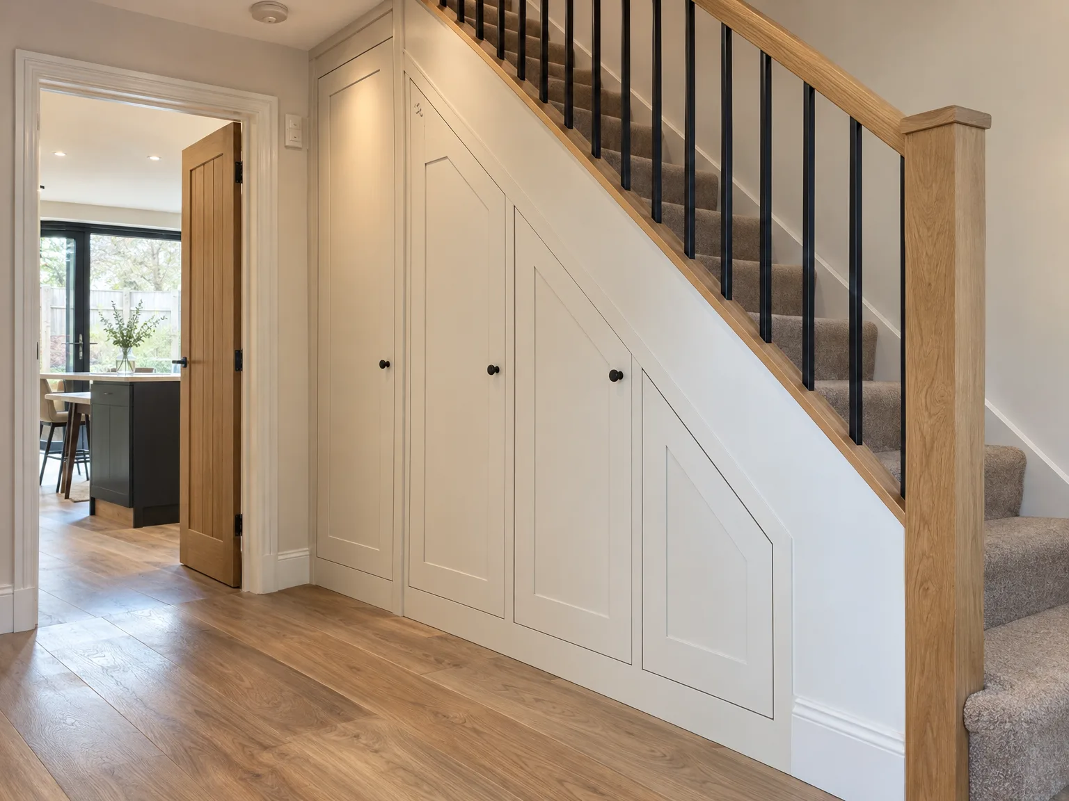 Built-in fitted storage cabinetry under modern staircase with wooden handrail, black spindles and grey carpet stairs in contemporary hallway